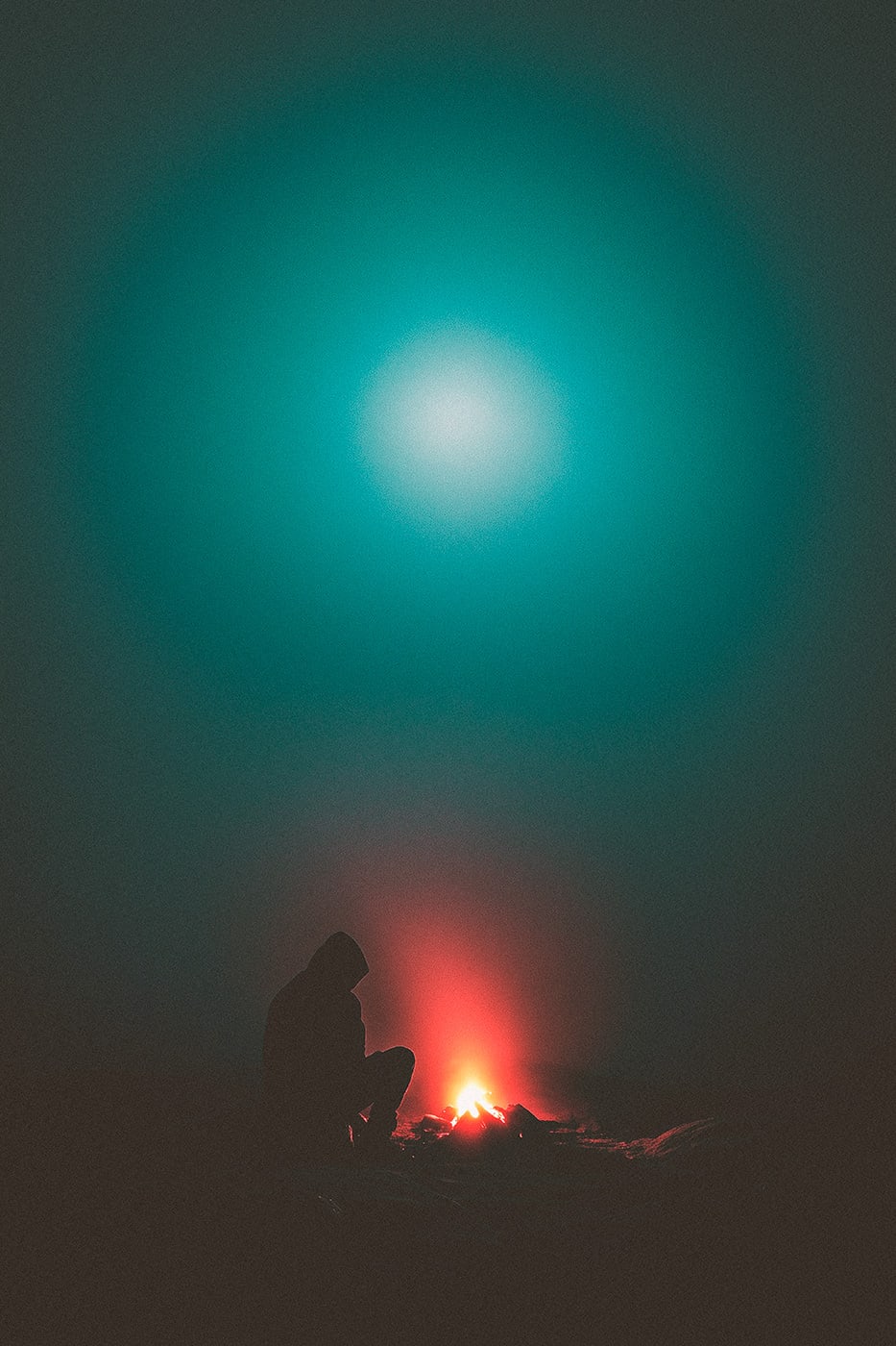 A mysterious hooded person sitting at a campfire in a dark, foggy environment. A soft, shimmering light is breaking through the fog. | Wall Art